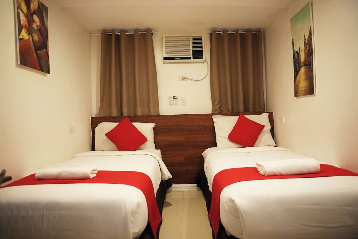 Quarto de hotel com duas camas de solteiro, cada uma com colcha vermelha e branca, cabeceira de madeira e janela com cortinas.