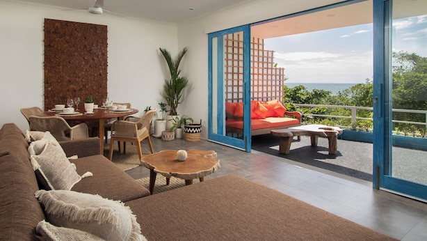 Een moderne woonkamer met een bank, salontafel en een eethoek. Er is een schuifdeur naar een balkon met uitzicht op de oceaan.