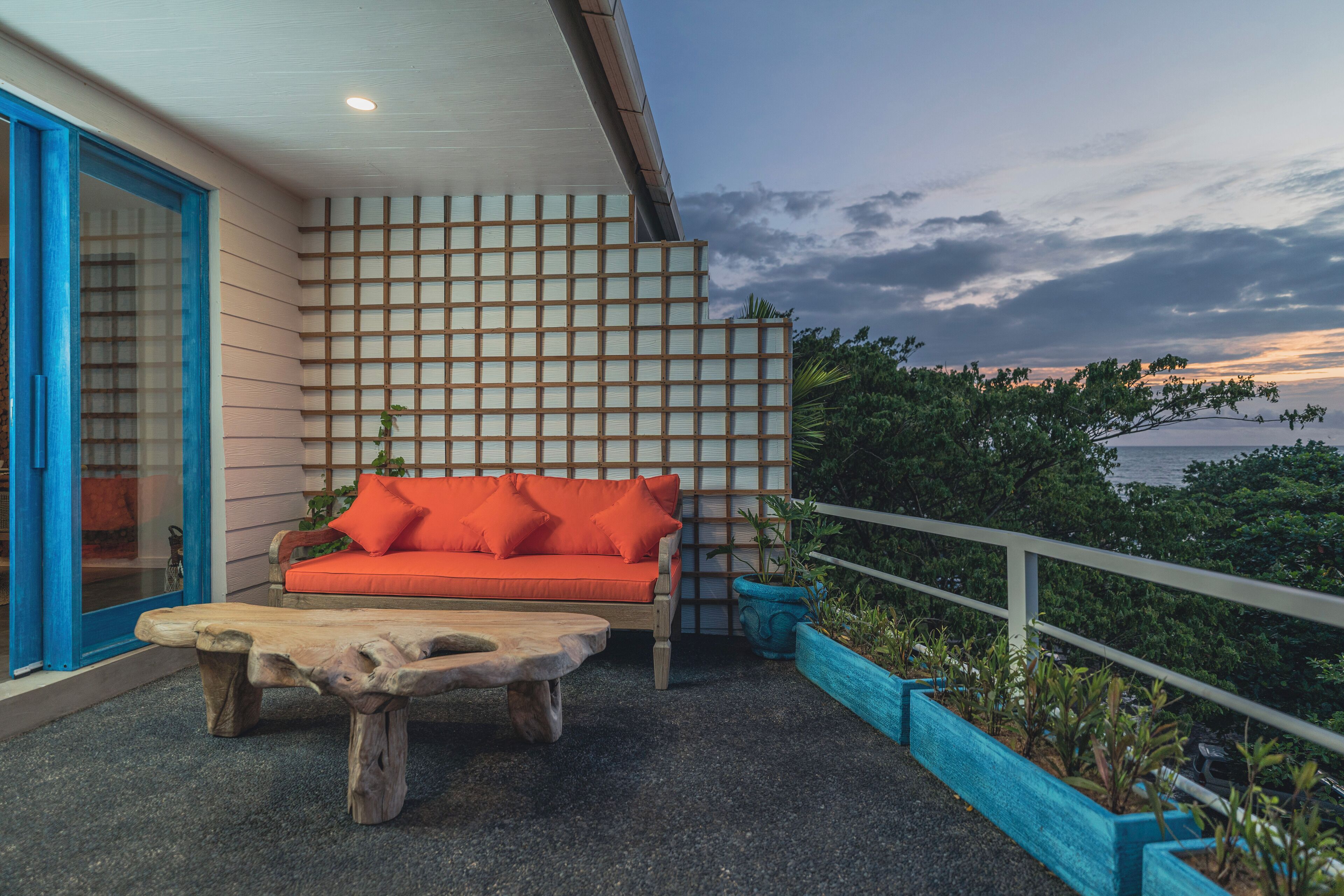 An outdoor seating area with a wooden table and orange cushions, a blue-framed door, and a view of the sea.