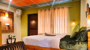 Ein Zimmer mit einem Bett, einer Sitzbank, einem kleinen Tisch mit Wasserkocher und einem Wandventilator.