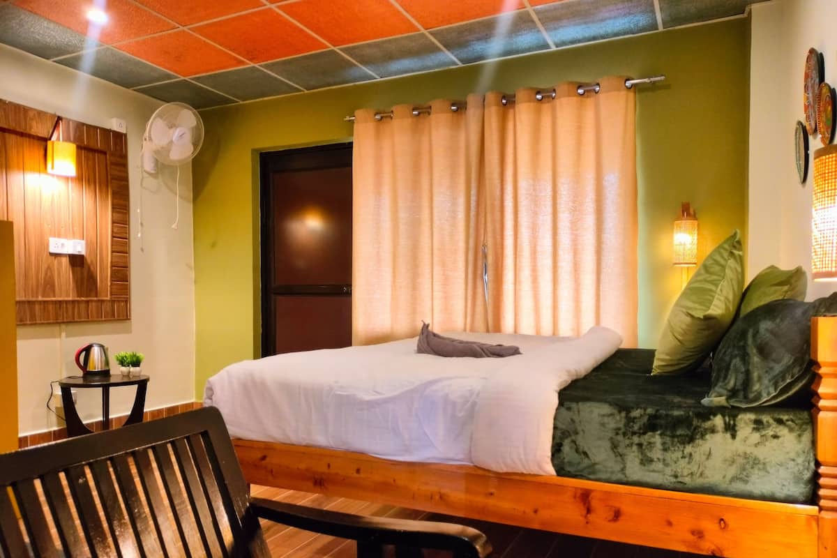 A room with a bed, a bench, a small table with a kettle, and a wall-mounted fan.