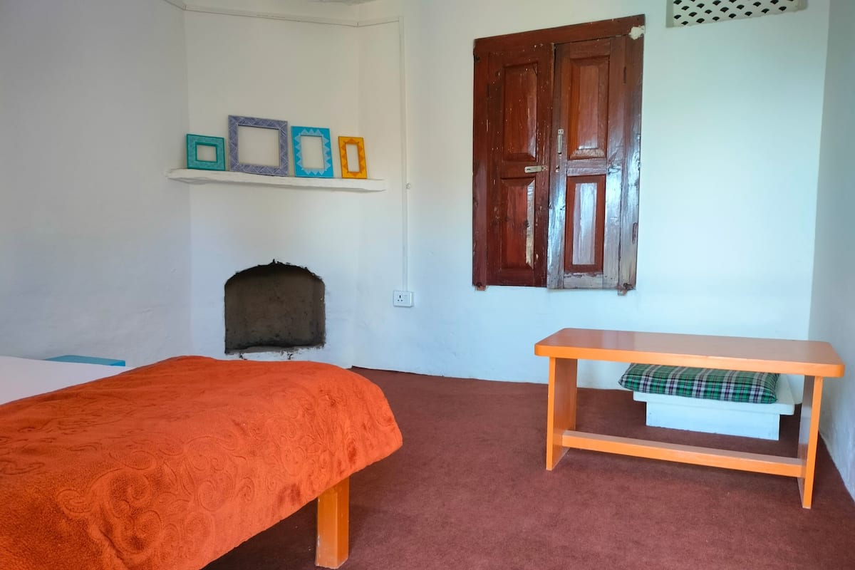 A room with a bed, a small table with a cushion, a shelf with picture frames, and a wooden door.