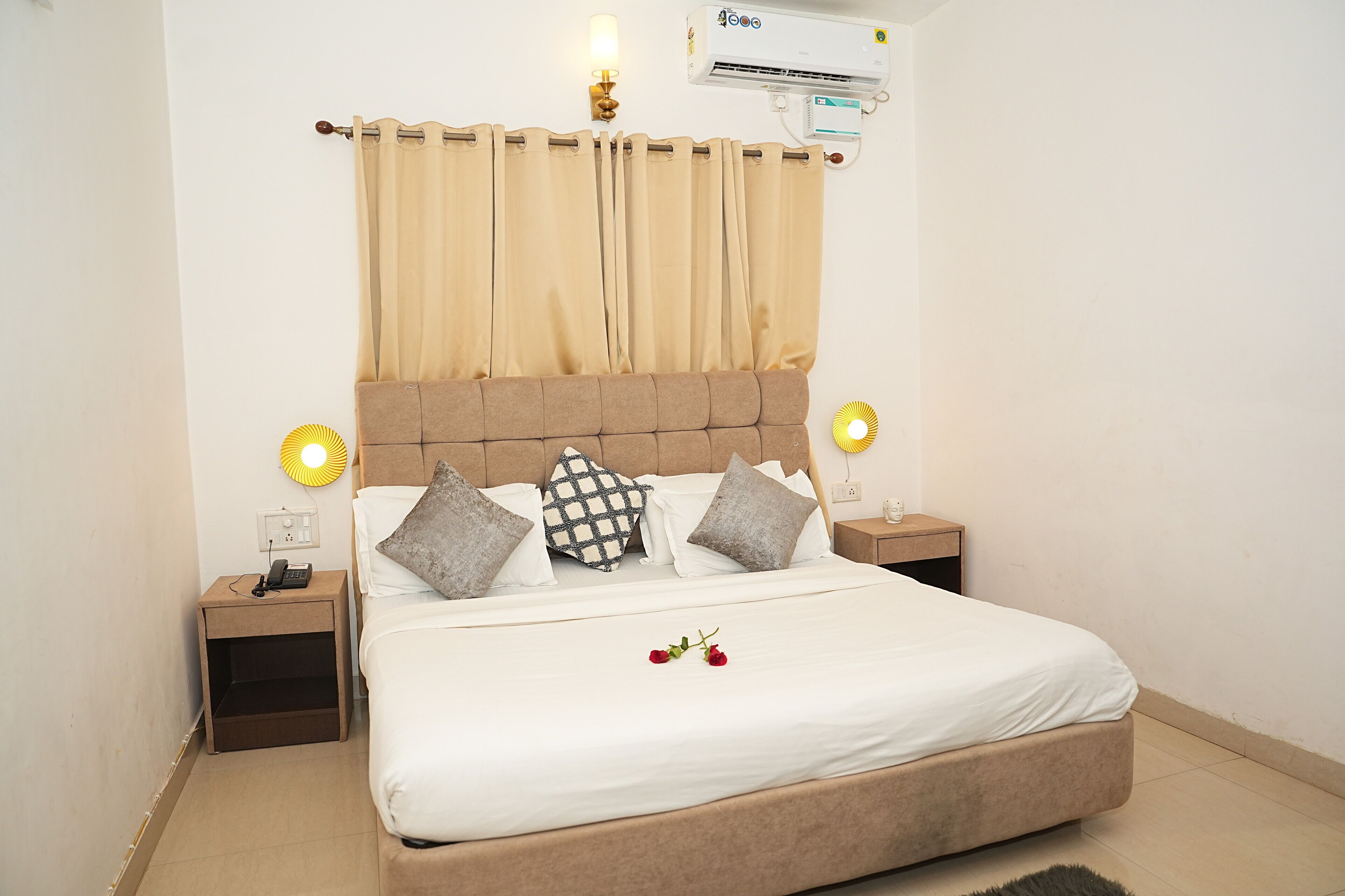Una cama bien tendida con un cabecero, dos mesitas de noche con lámparas, un aire acondicionado de pared y una cortina.