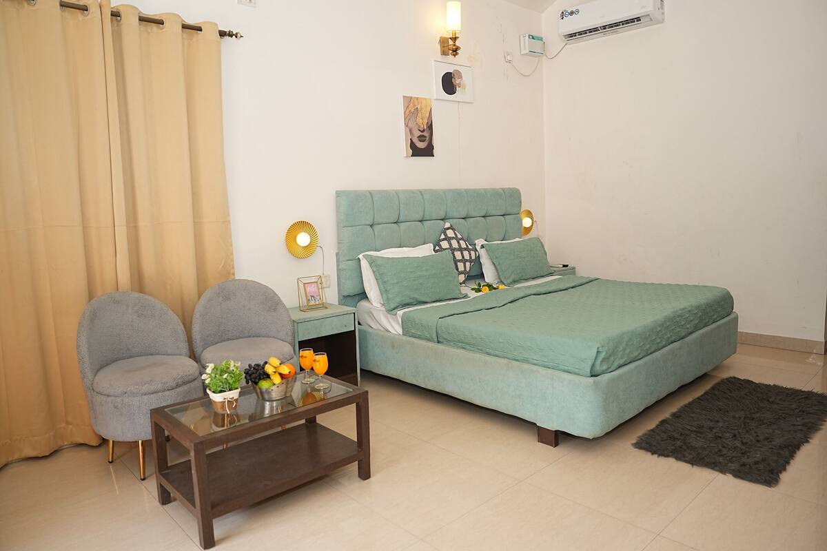 Une chambre à coucher avec un lit, deux chaises, une petite table avec une plante et des boissons, et un tapis.
