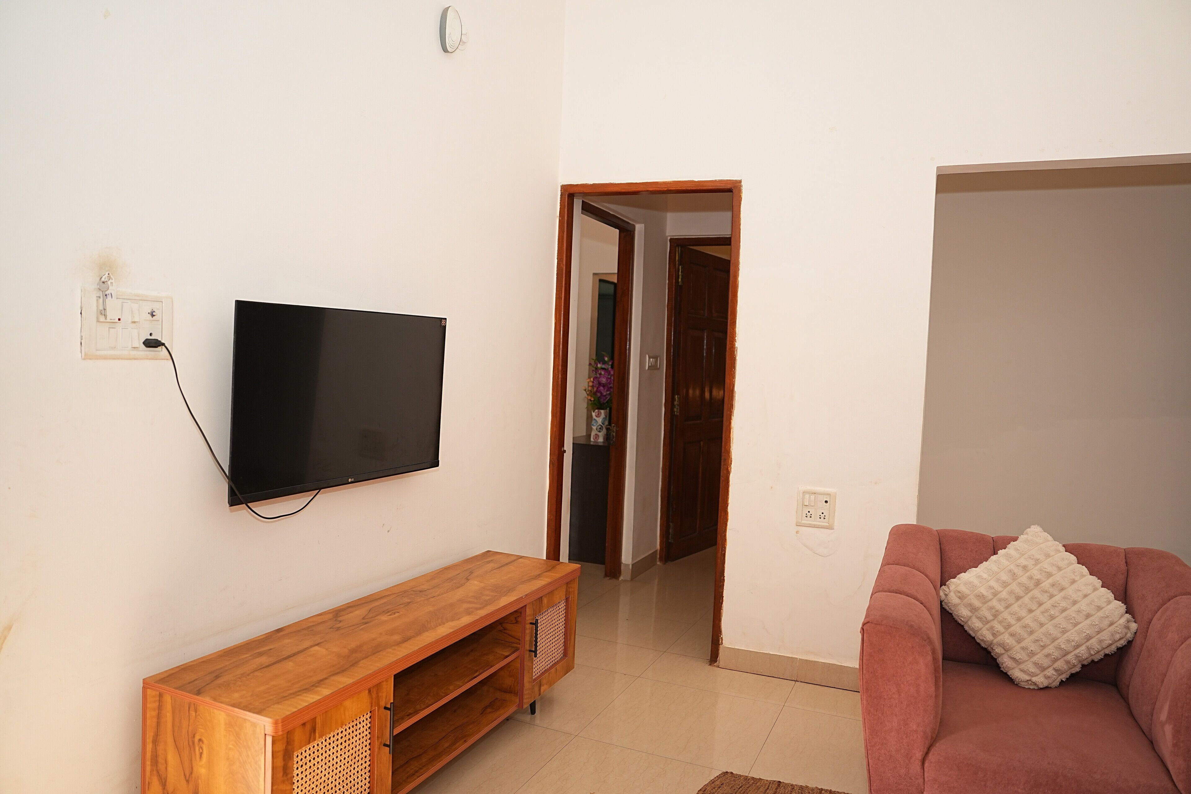Ein Wohnzimmer mit Wand-Fernseher, einem Holzhochentisch, einem rosafarbenen Sofa mit Zierkissen und einer Tür zu einem anderen Raum.