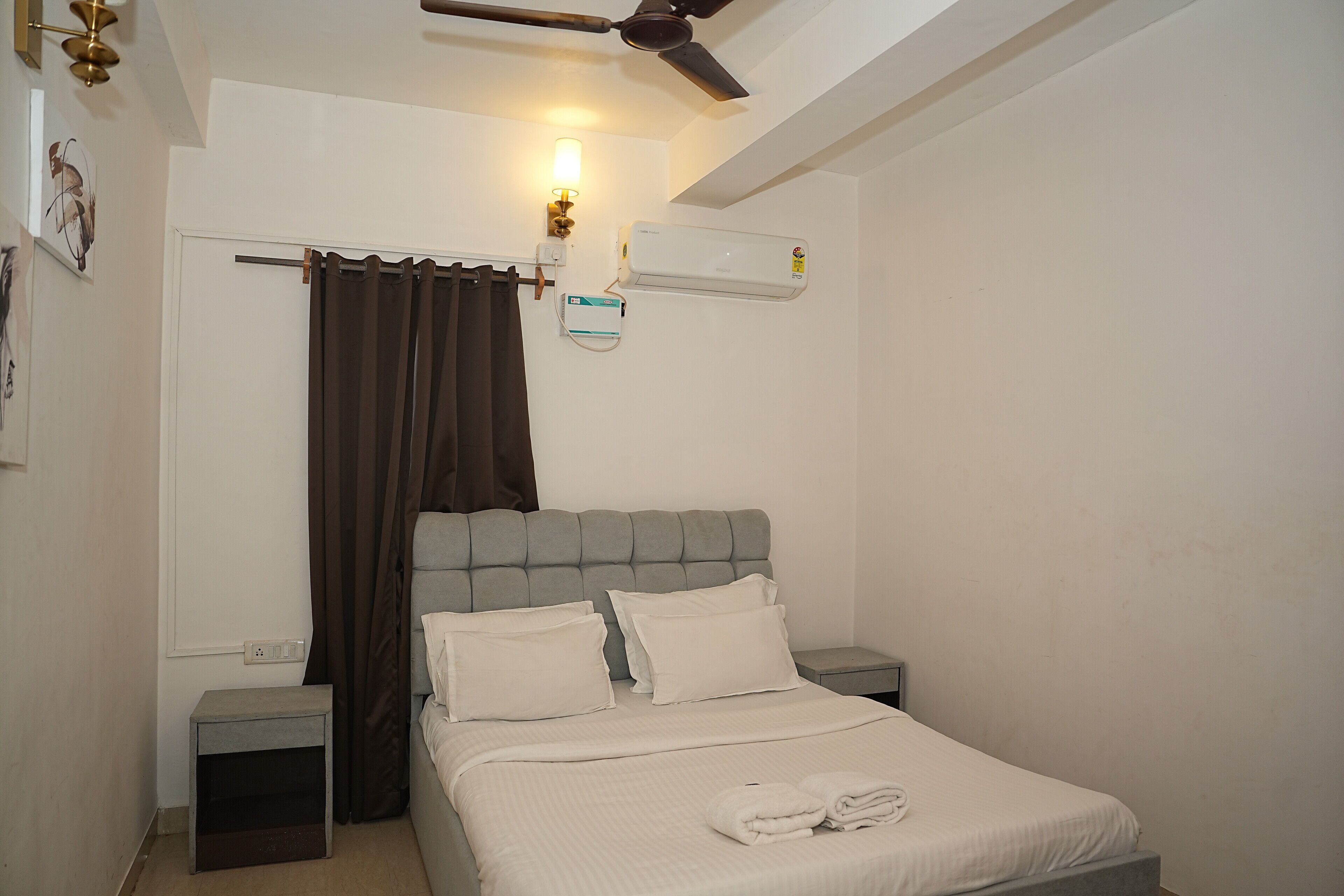 A neatly made bed with a grey headboard, a bedside table, a wall-mounted air conditioner, and a ceiling fan.