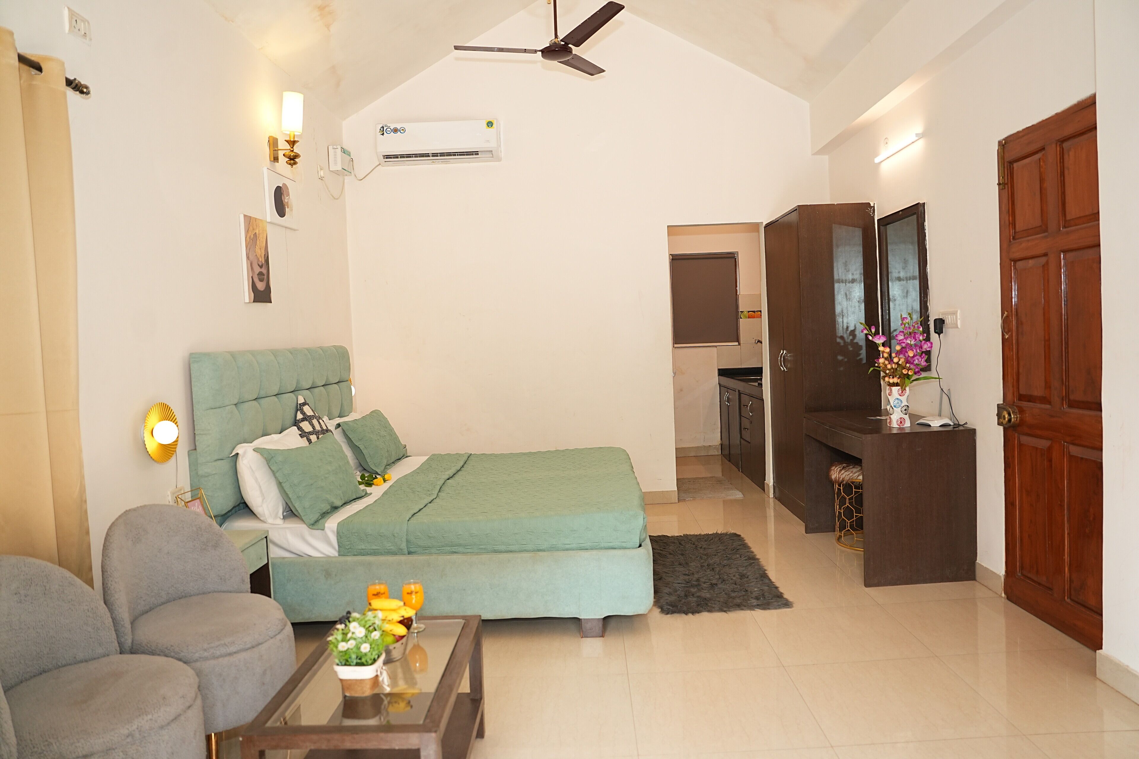 A well-lit, modern living room with a sofa, a bed with green bedding, a coffee table with flowers, and a ceiling fan.