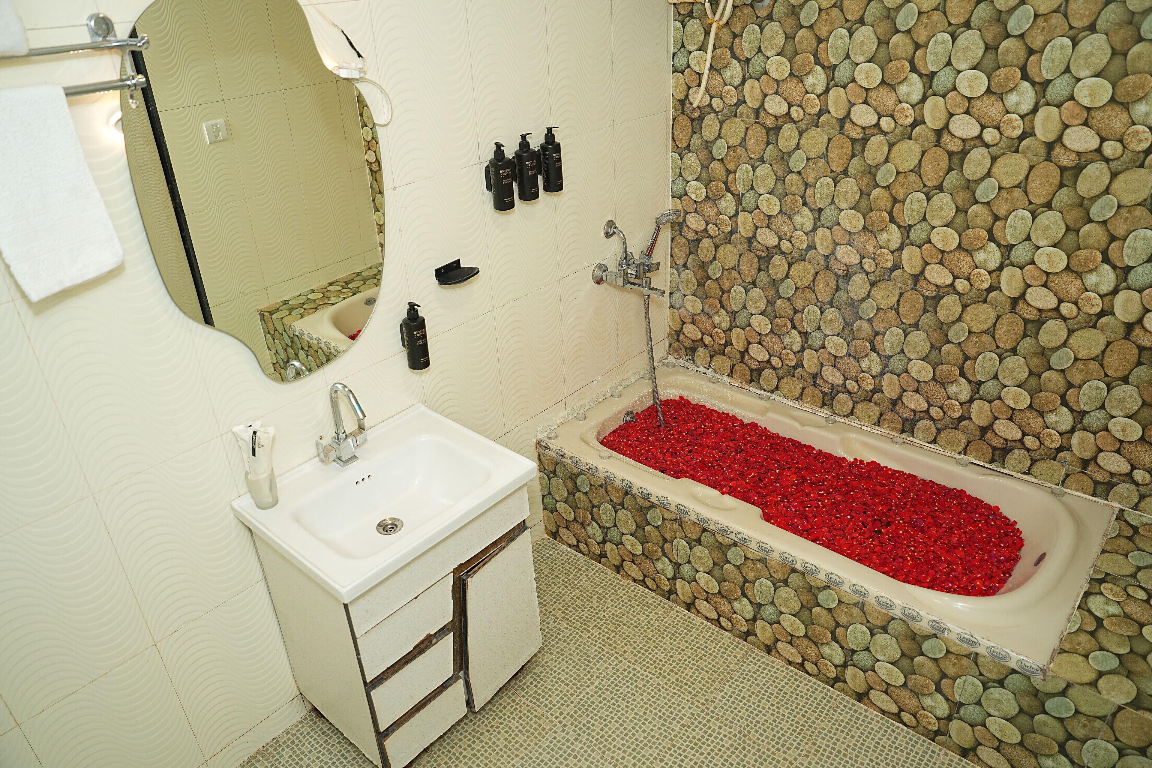 A bathroom with a sink, mirror, toiletries, and a bathtub with red pebbles.