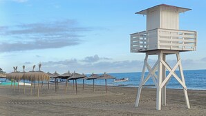 A beach with a lifeguard tower, thatched umbrellas, and a boat on the water.