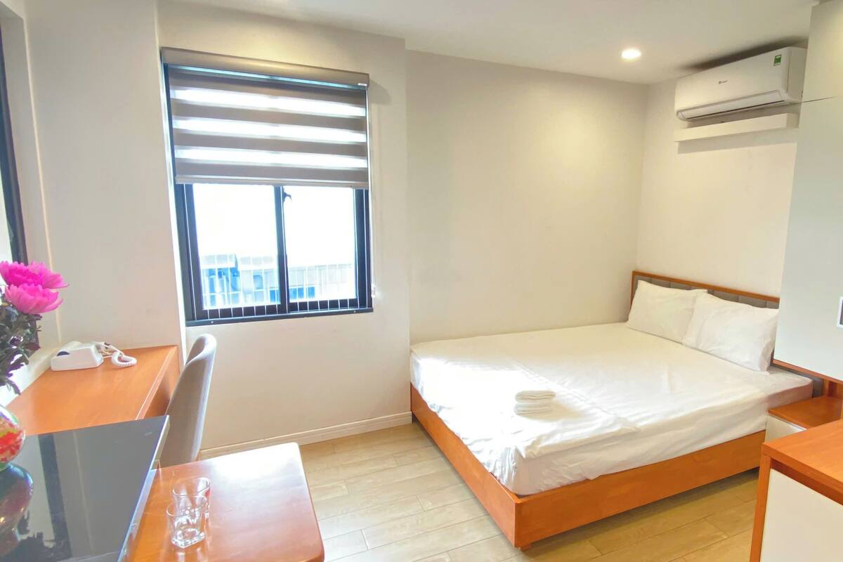 Une chambre à coucher comprenant un lit, un bureau, une chaise, une fenêtre avec des stores, un appareil de climatisation et une petite table sur laquelle est posé un vase de fleurs.