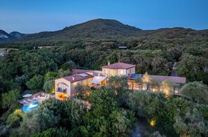 Una casa grande de varios pisos rodeada de un bosque frondoso, con piscina y un espacio cubierto al aire libre.