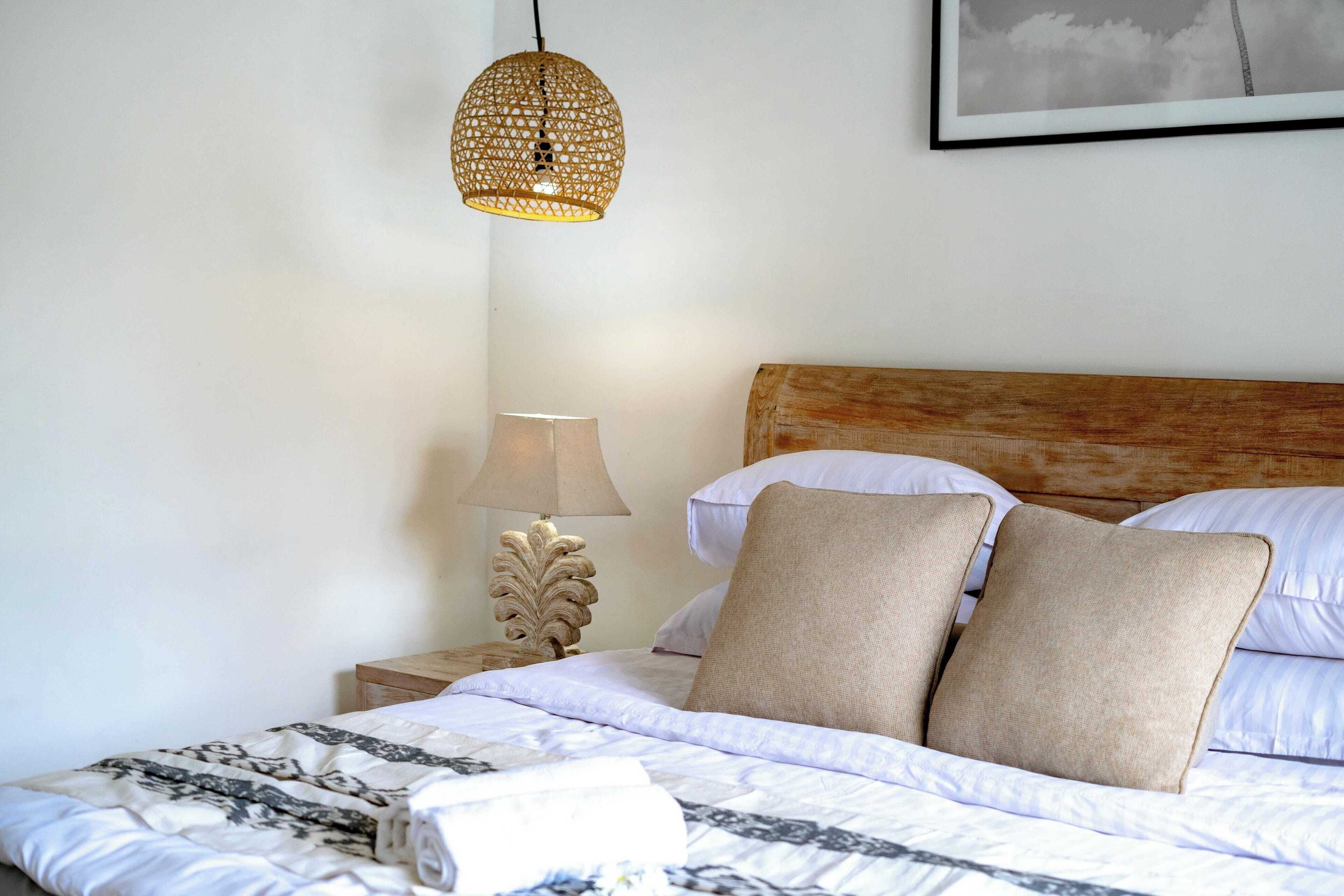 Een keurig opgemaakt bed met een bedspreide met dessin, twee beige kussens, een houten hoofdbord, een bedlampje en een geweven hanglamp.