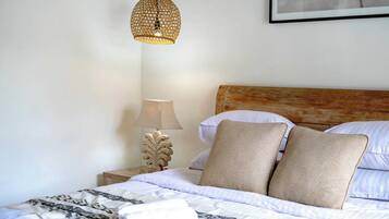 Een keurig opgemaakt bed met een bedspreide met dessin, twee beige kussens, een houten hoofdbord, een bedlampje en een geweven hanglamp.