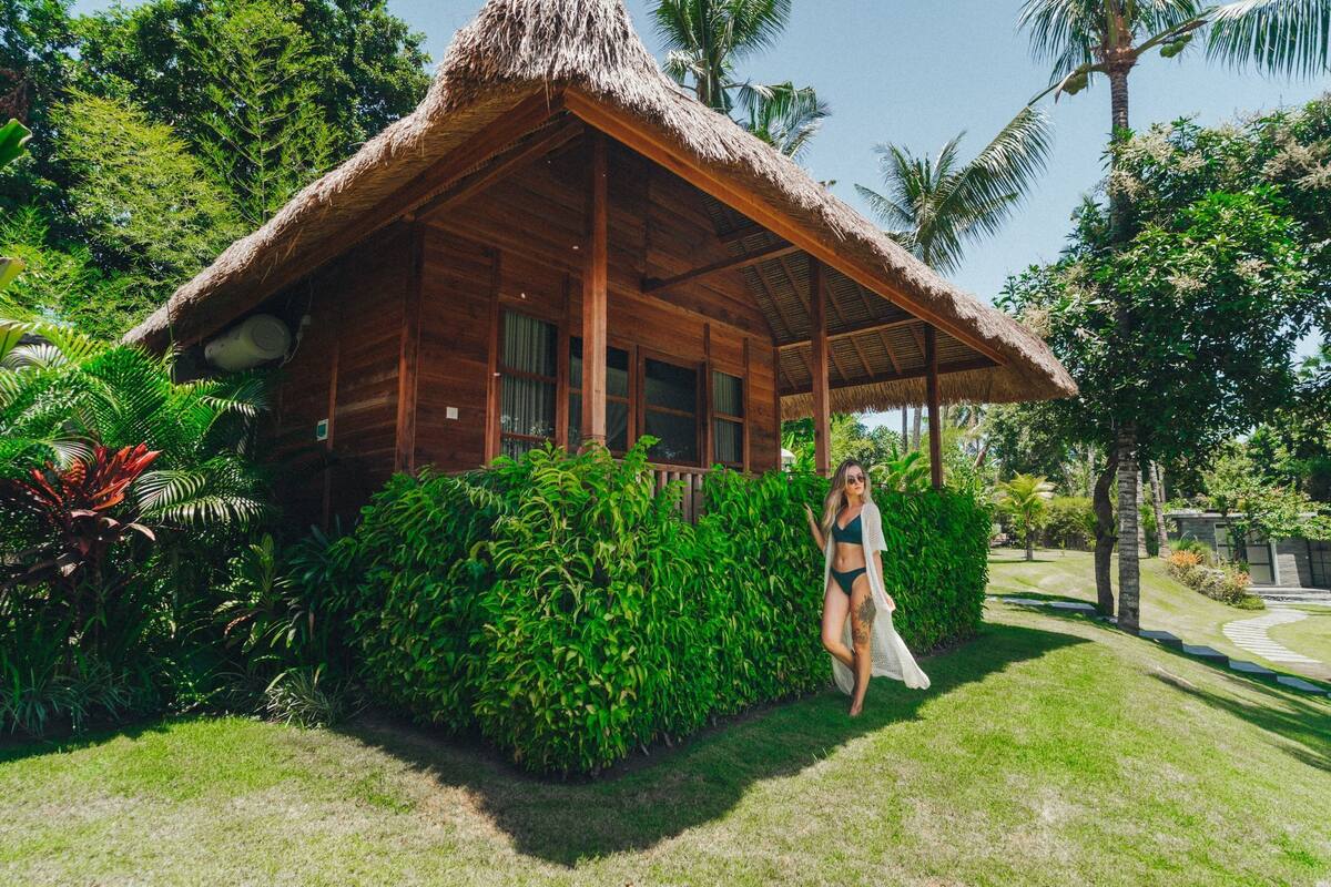 A wooden cabin with a thatched roof surrounded by lush greenery.
