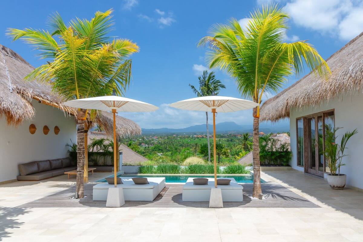 A pool area with two lounge chairs and umbrellas, surrounded by palm trees and a view of a mountainous landscape.