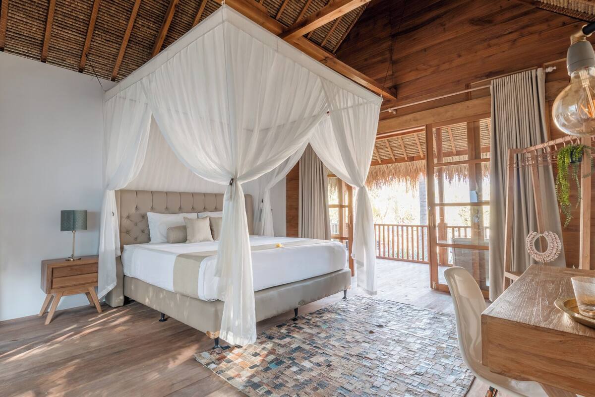 A bedroom with a four-poster bed, wooden flooring, and a large window with curtains.