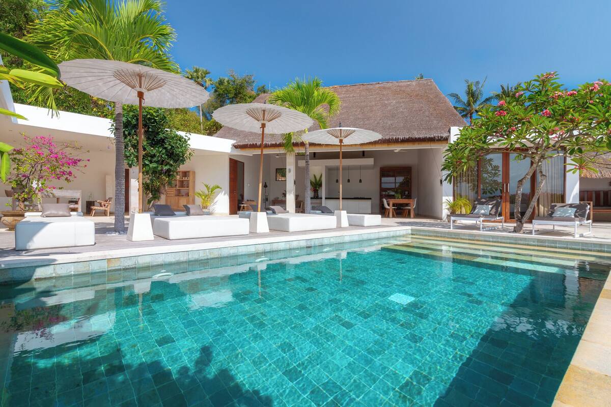 A clear blue pool with white lounge chairs and umbrellas, surrounded by greenery and a thatched-roof structure.