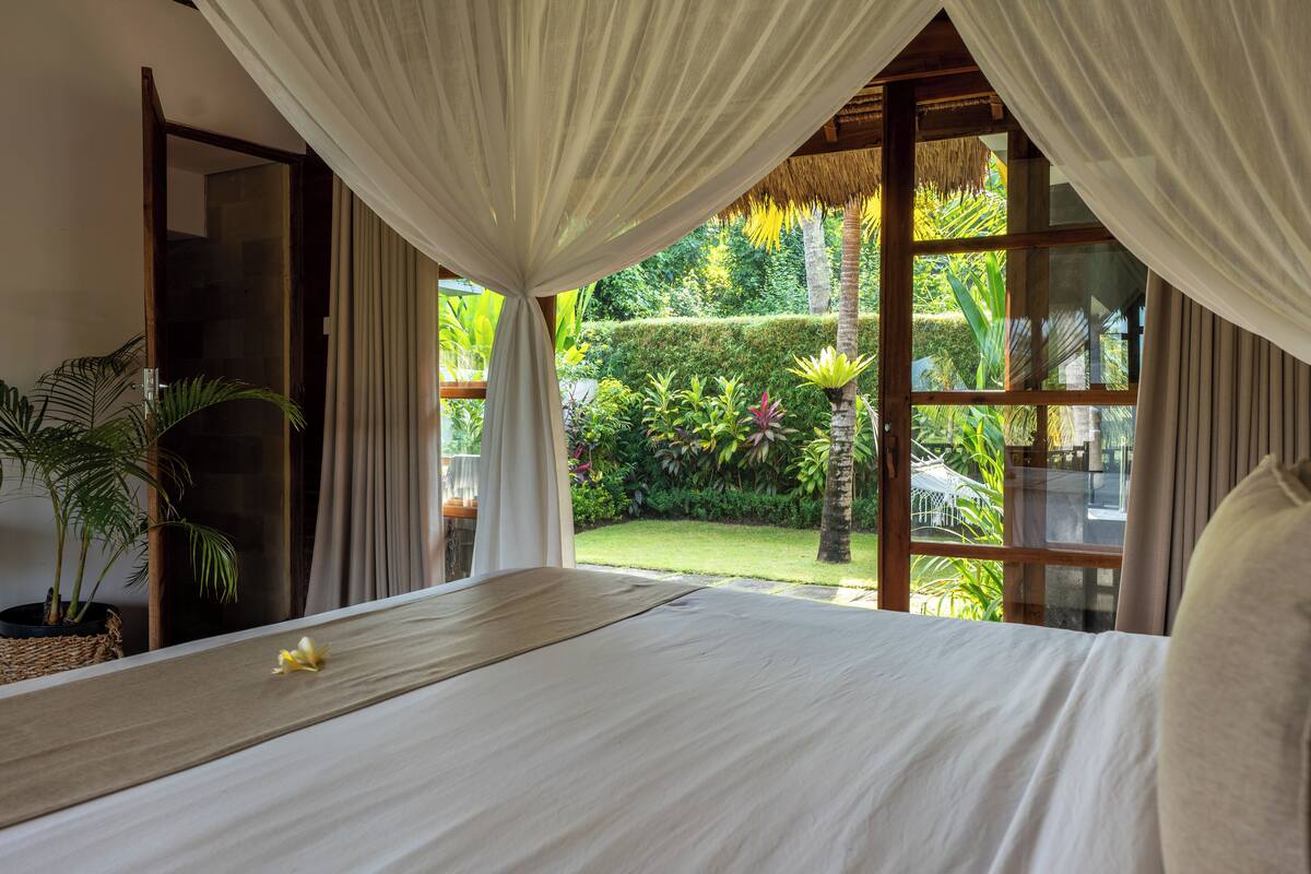 A bedroom with a large bed, a view of a lush garden, and a thatched roof structure.