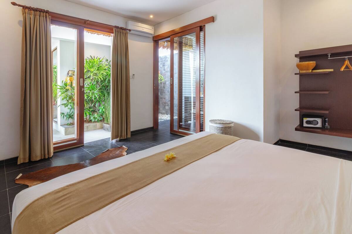 A hotel room with a bed, a small shelf with a radio, and a view of a garden through the sliding glass door.