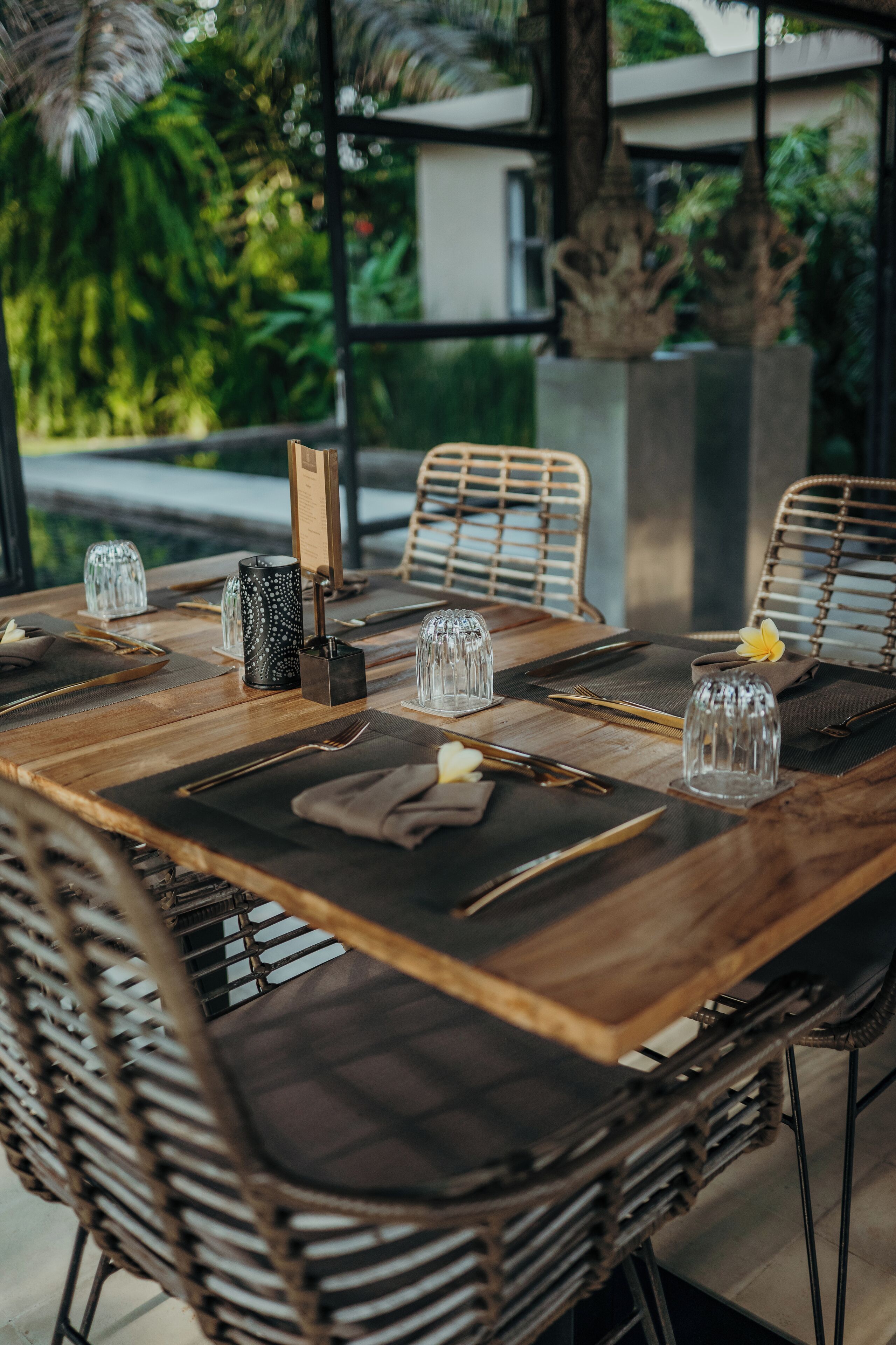 A wooden dining table set with glassware, cutlery, and napkins, surrounded by wicker chairs.