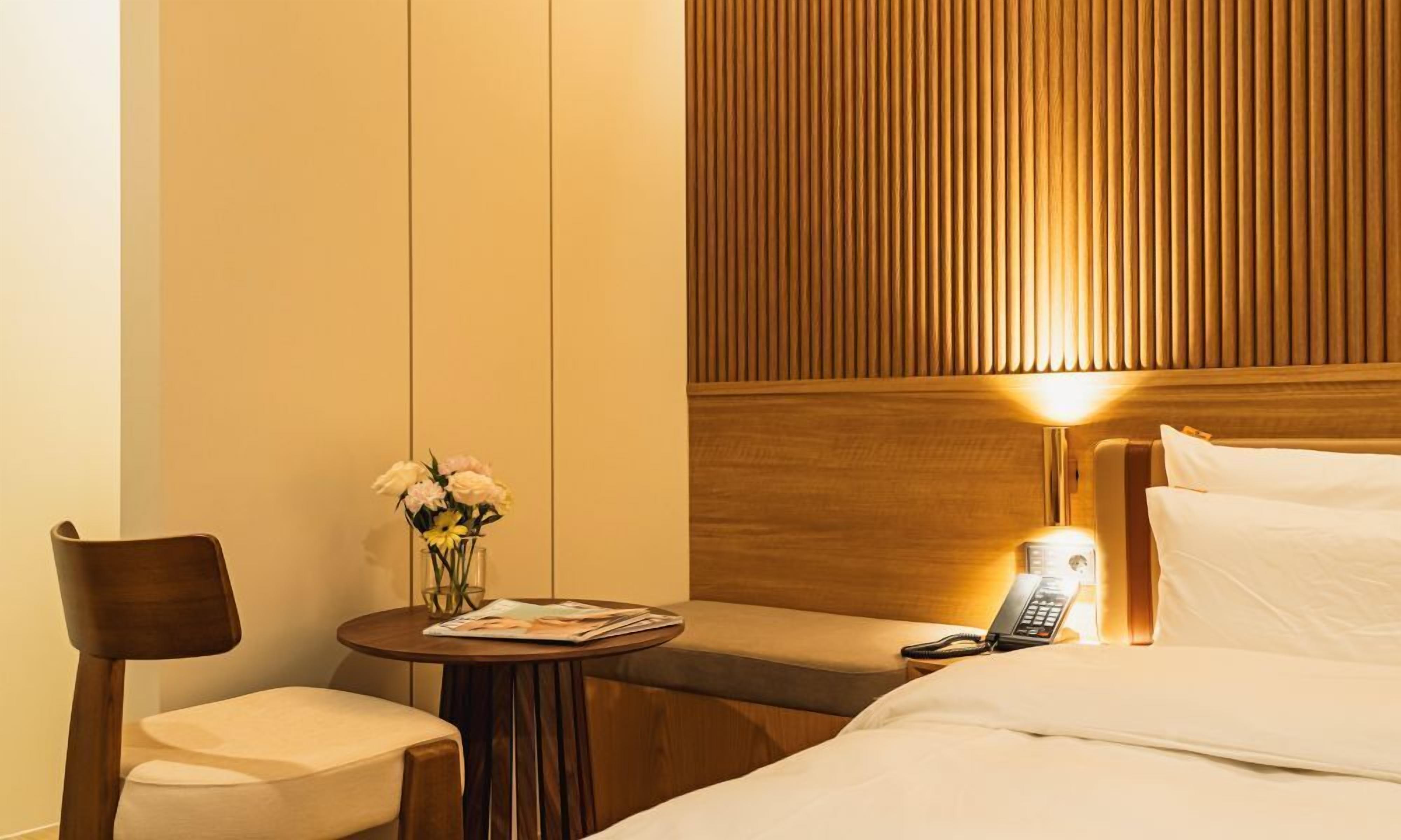 A hotel room with a bed, a bedside table with a lamp, a chair, and a small table with a vase of flowers.