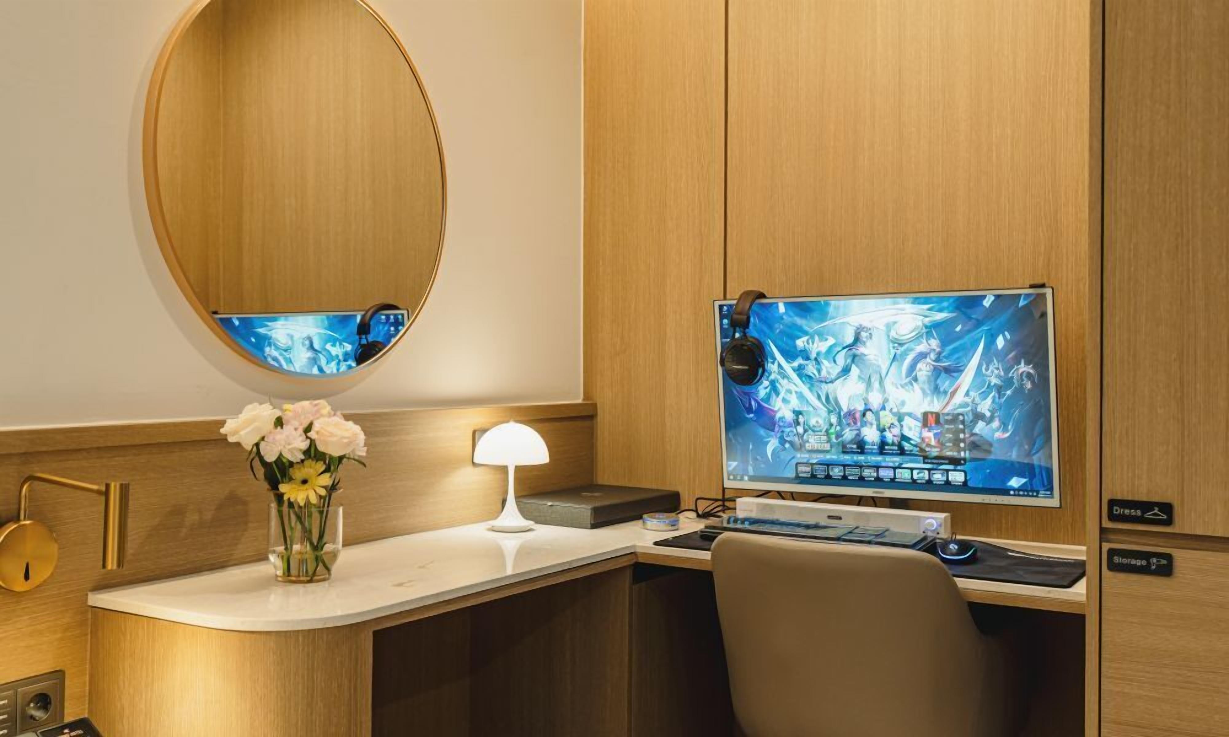 A hotel room with a wooden desk, a computer monitor, a lamp, and a vase of flowers.