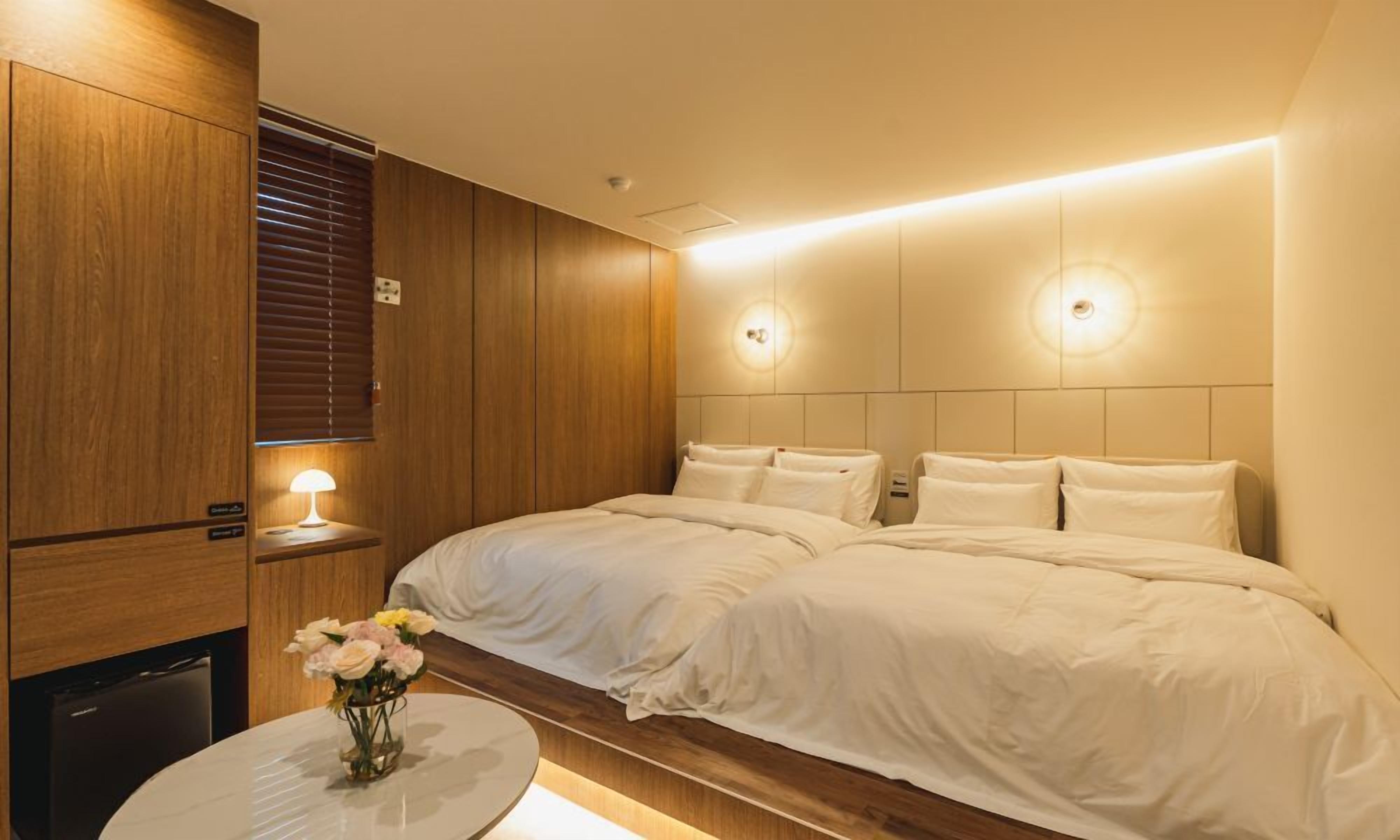A hotel room with two beds, wooden paneling, a small table with a lamp, and a vase of flowers.