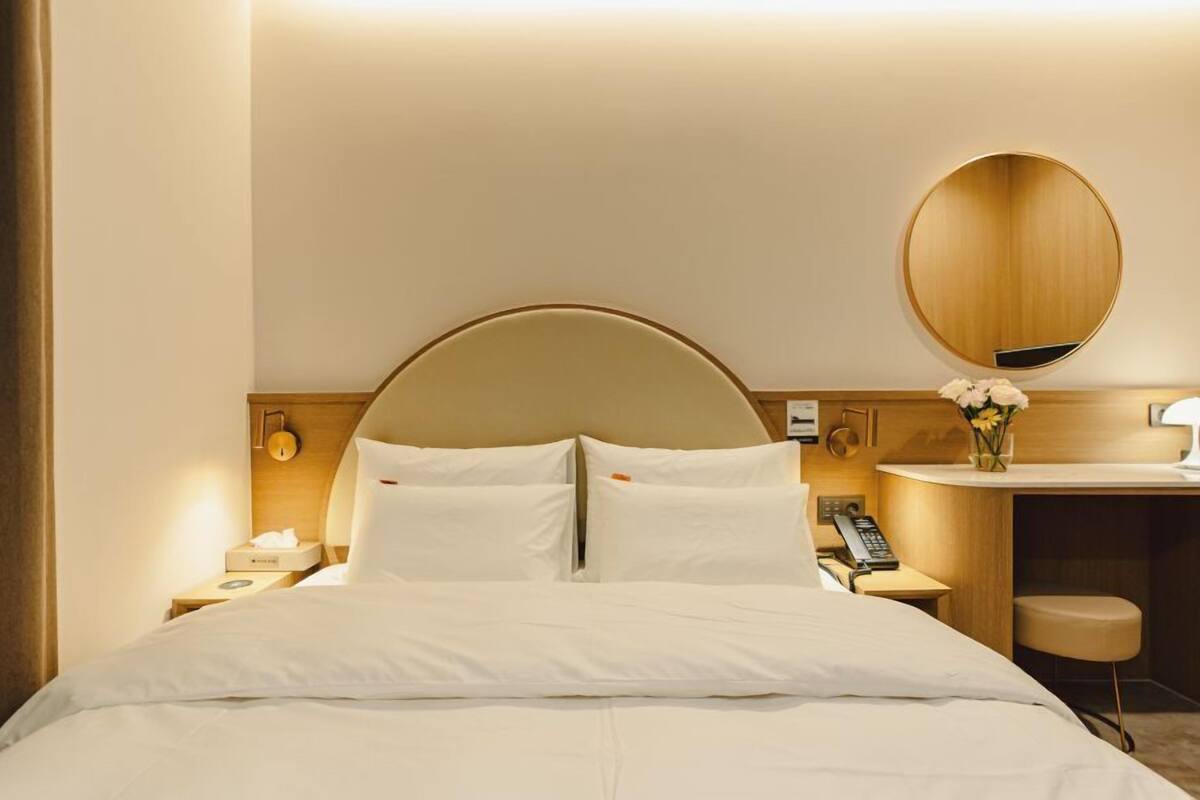 A hotel room with a large bed, a round mirror, a bedside table with a lamp, and a small stool.