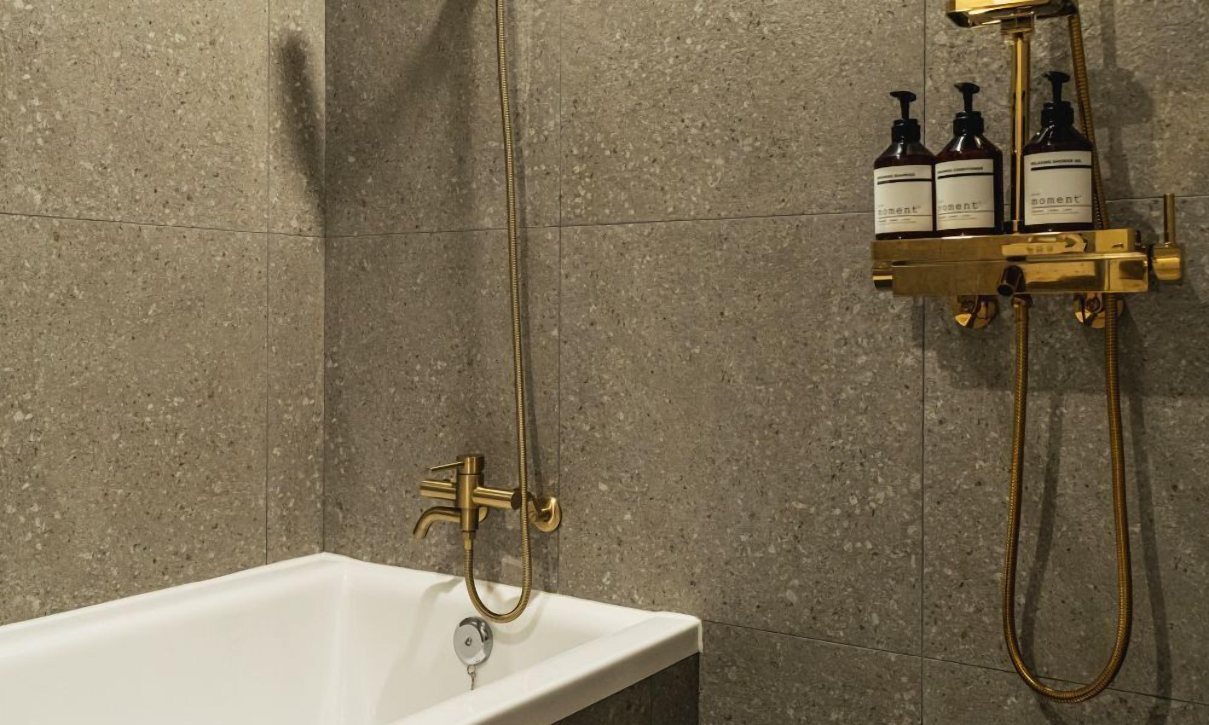 Un baño con una bañera blanca, un cabezal de ducha dorado y dos botellas de jabón de manos Moment sobre un estante dorado.