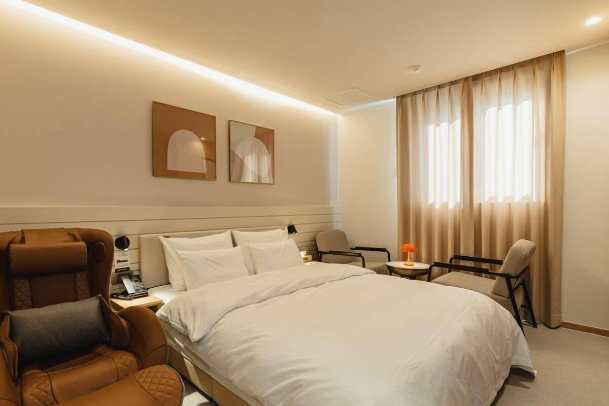 A hotel room with a bed, a brown leather chair, a small table with a lamp, and a window with curtains.