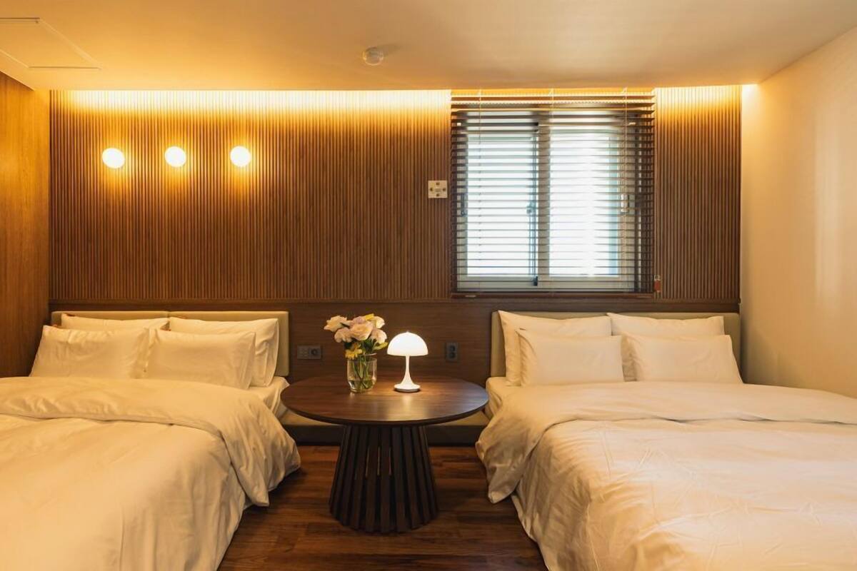 A hotel room with two beds, a round table with a lamp, and a window with blinds.