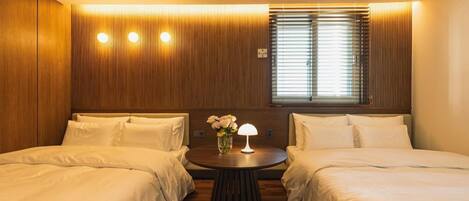 Una habitación de hotel con dos camas, una mesa redonda con lámpara y una ventana con persianas.