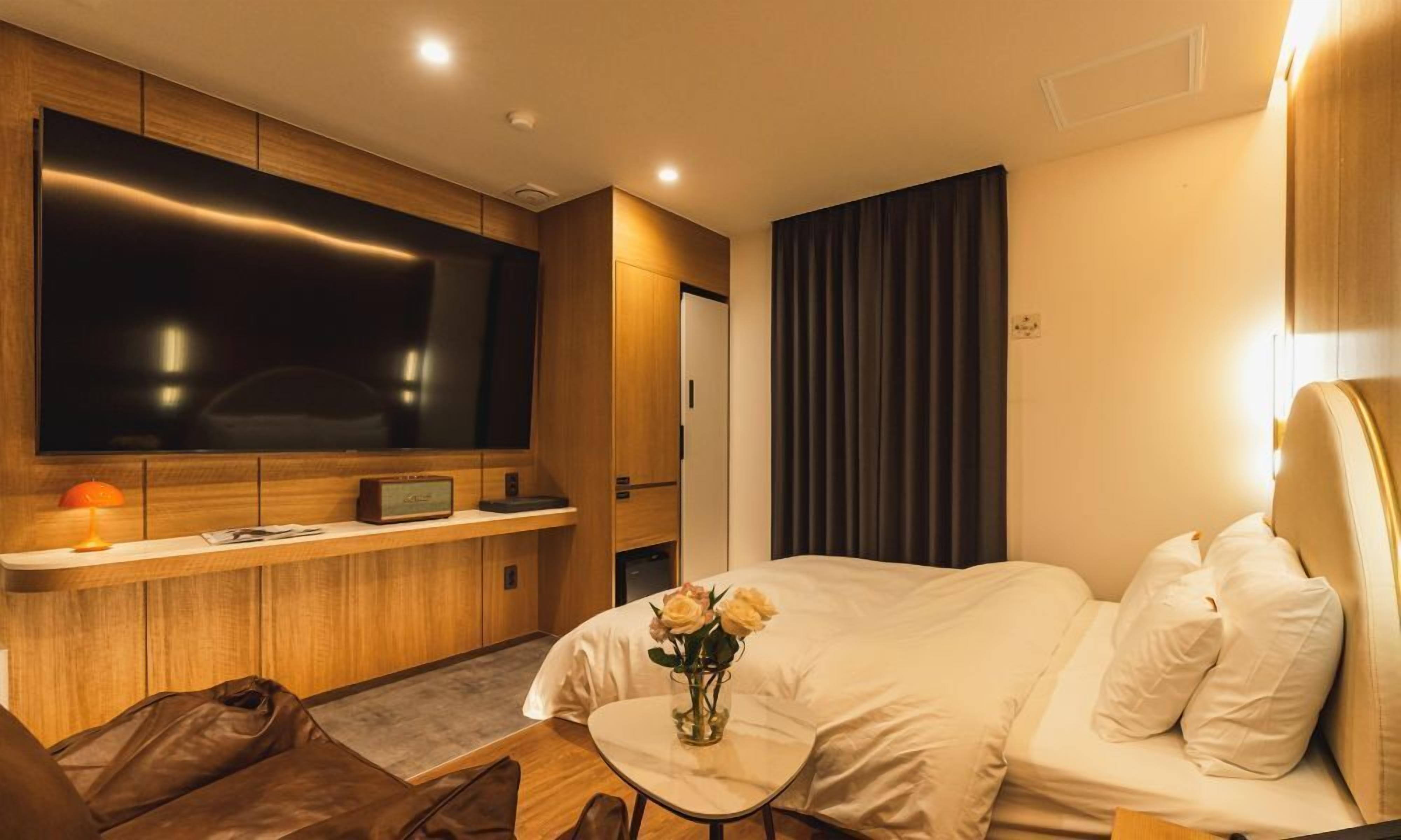 A modern hotel room with a large flat-screen TV, a small table with a vase of flowers, a bed with white linens, and a seating area with a brown leather couch.