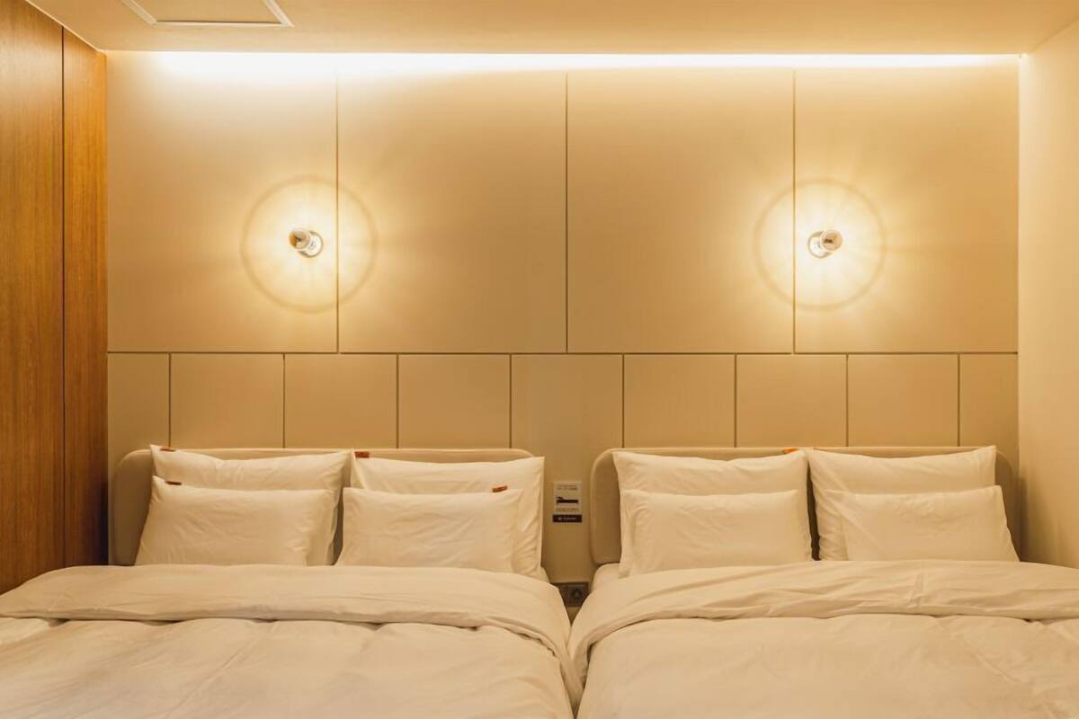 A hotel room with two beds, a wooden panel wall, and wall-mounted lighting.