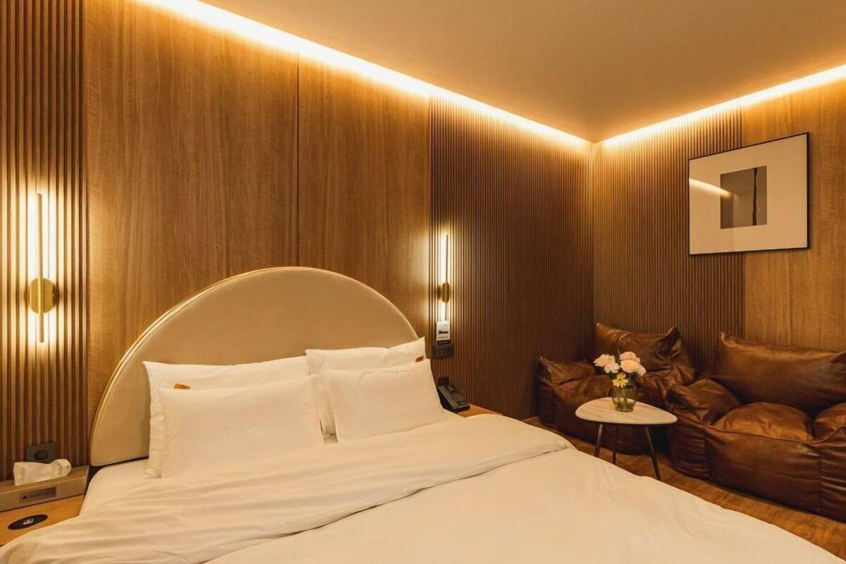 A modern hotel room with a large bed, a brown leather sofa, a small table with a vase of flowers, and wall-mounted lights.