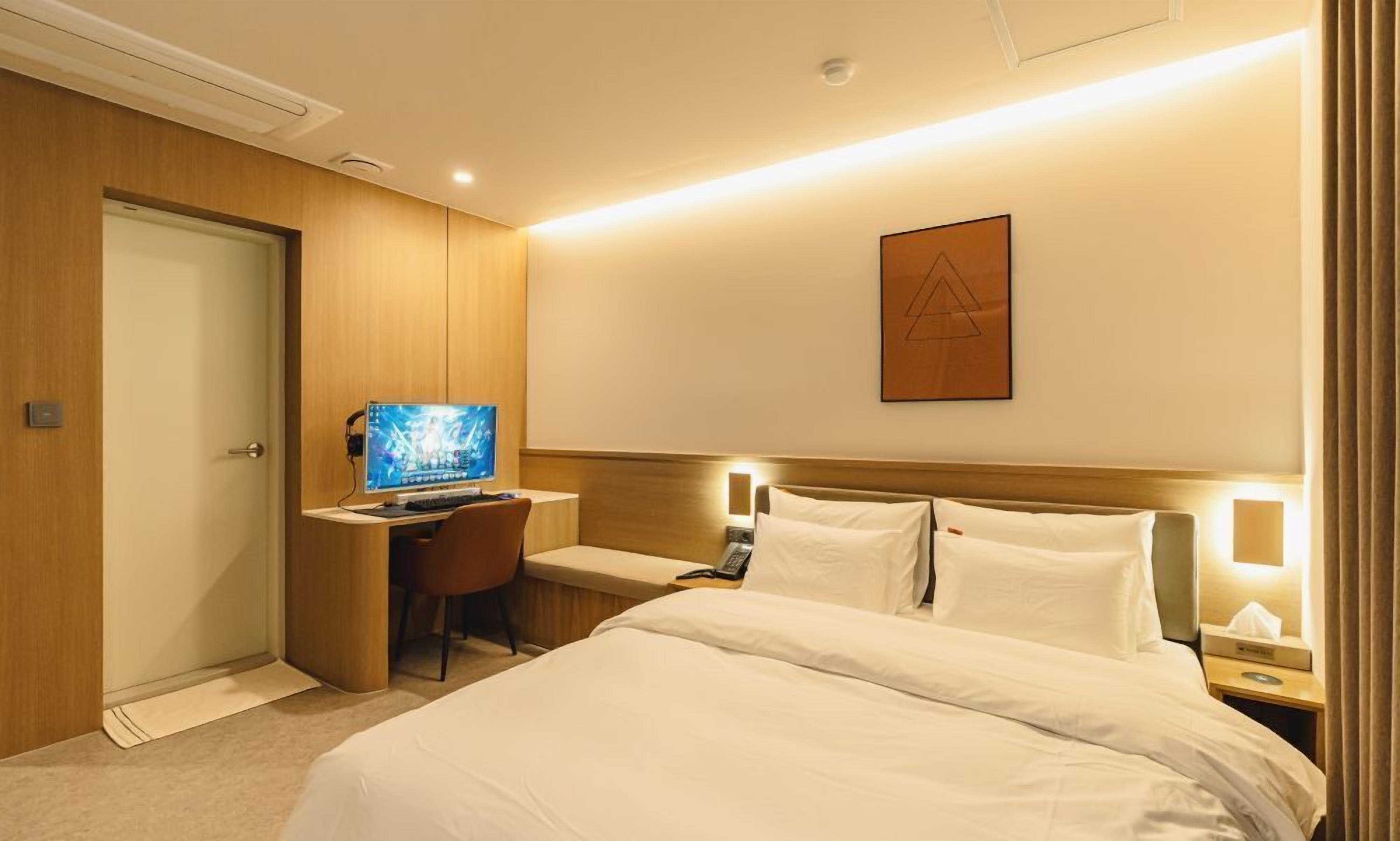 Habitación de hotel con cama, escritorio con computadora, silla y mesita de noche con lámpara.