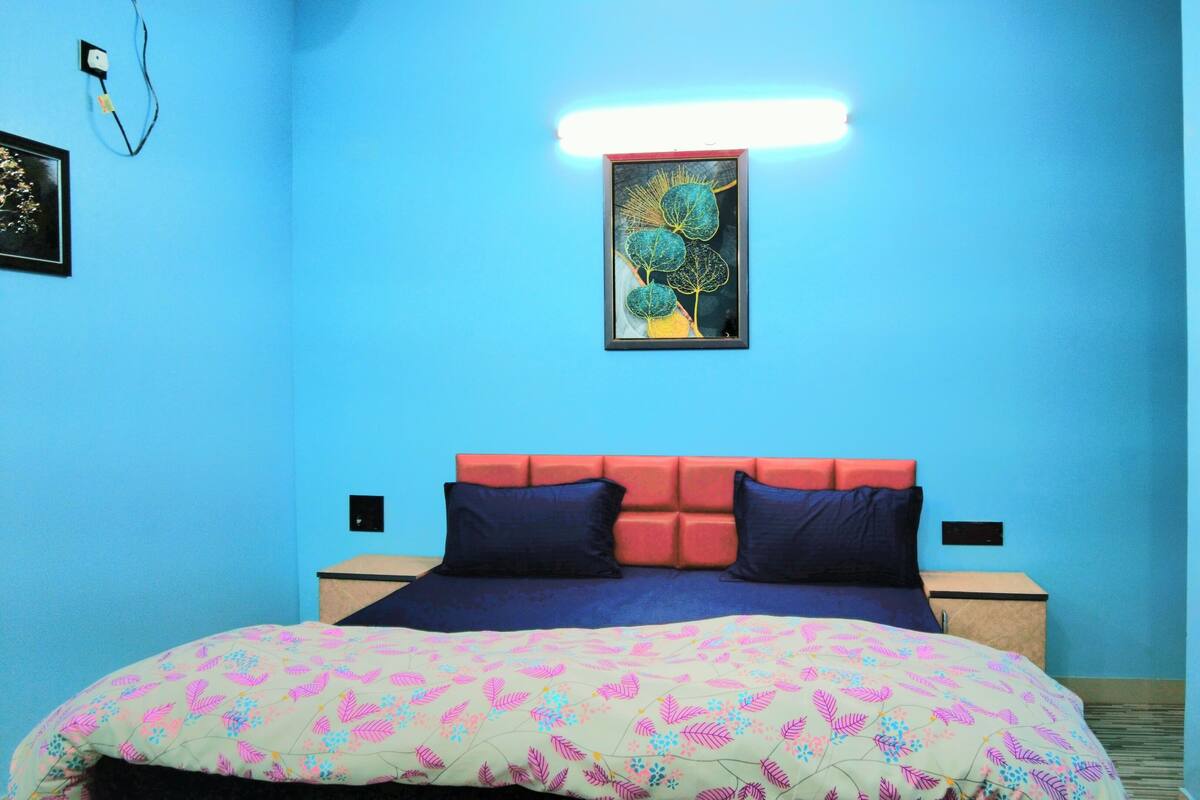 A room with a blue wall, a bed with a colorful floral bedspread, a framed artwork above the bed, and a bedside table with a lamp.