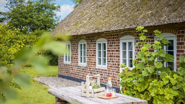 A rustic house with a thatched roof, brick walls, and a wooden table with a basket and glass bottles.
