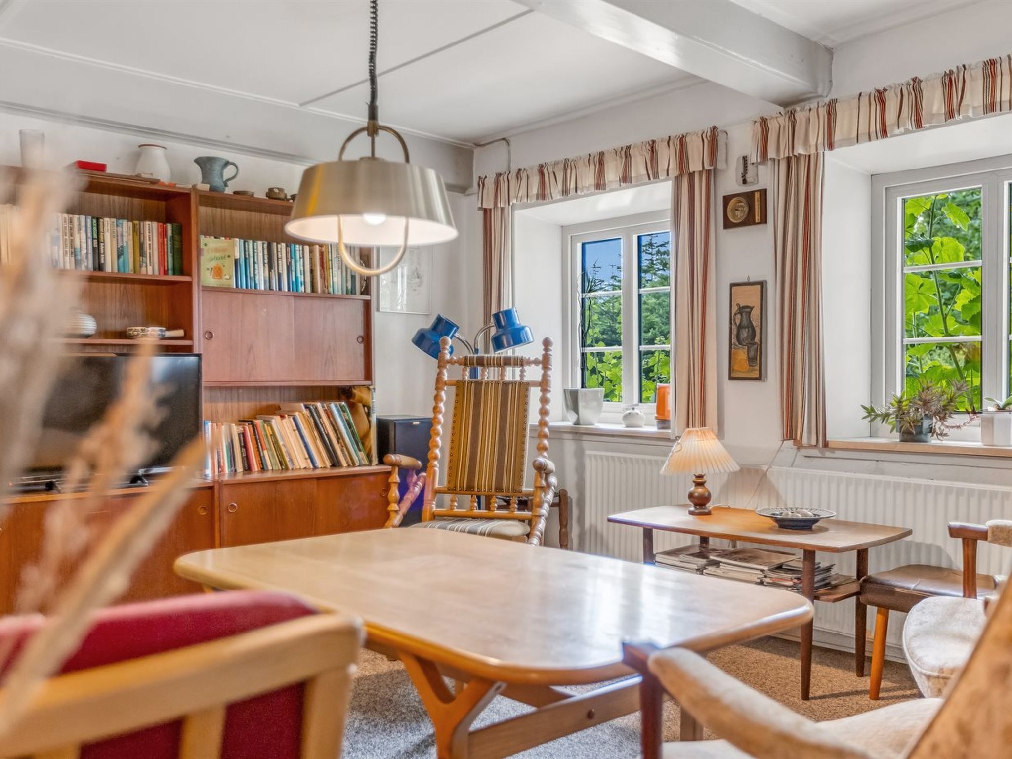 Ein gemütliches Zimmer mit einer Holzbücherwand, einem Lesesessel, einem Tisch und einem Fenster mit Blick auf Grünflächen.