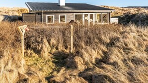 Una casa moderna con techo inclinado, ventanas grandes y una chimenea. Está rodeada de hierba alta y dunas.
