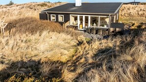 Una casa moderna con techo oscuro y ventanas amplias, rodeada de hierba alta y dunas.