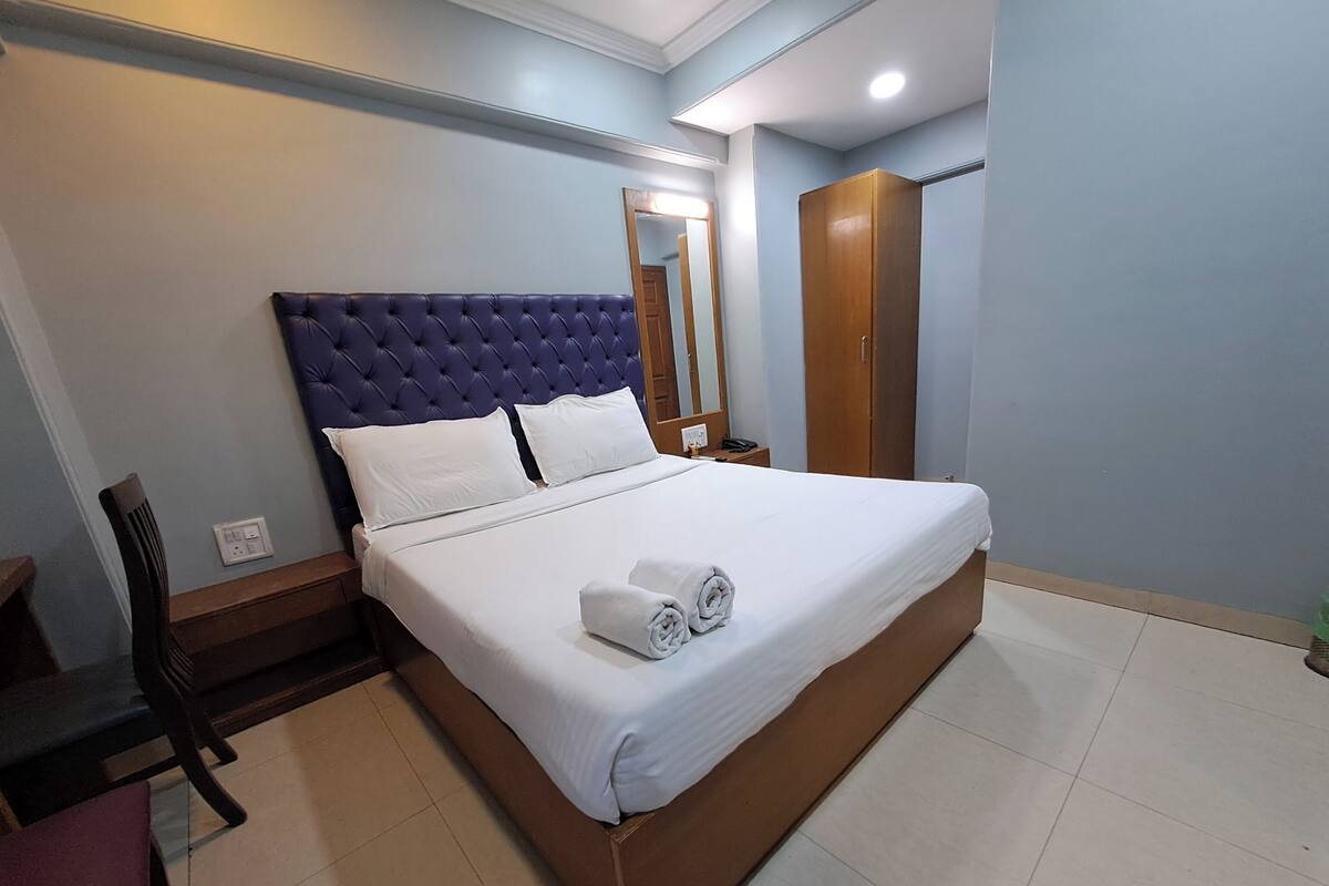 A neatly made bed with white linens and rolled towels, a wooden wardrobe, a chair, and a nightstand with a phone.