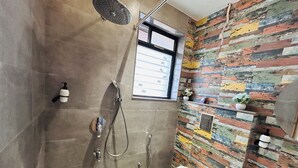 A modern bathroom with a shower, toilet, and sink. The shower has a rain showerhead and a handheld showerhead. The toilet has a white lid with a pink label. The sink is white and has a soap dispenser. The walls are tiled with a mix of colors. - HOTEL MEENA AURUM INN (Darbhanga)