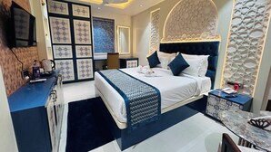 A modern hotel room with a large bed, a desk, and a chair. There is a ceiling fan, a TV, and a decorative wall panel. - HOTEL MEENA AURUM INN (Darbhanga)