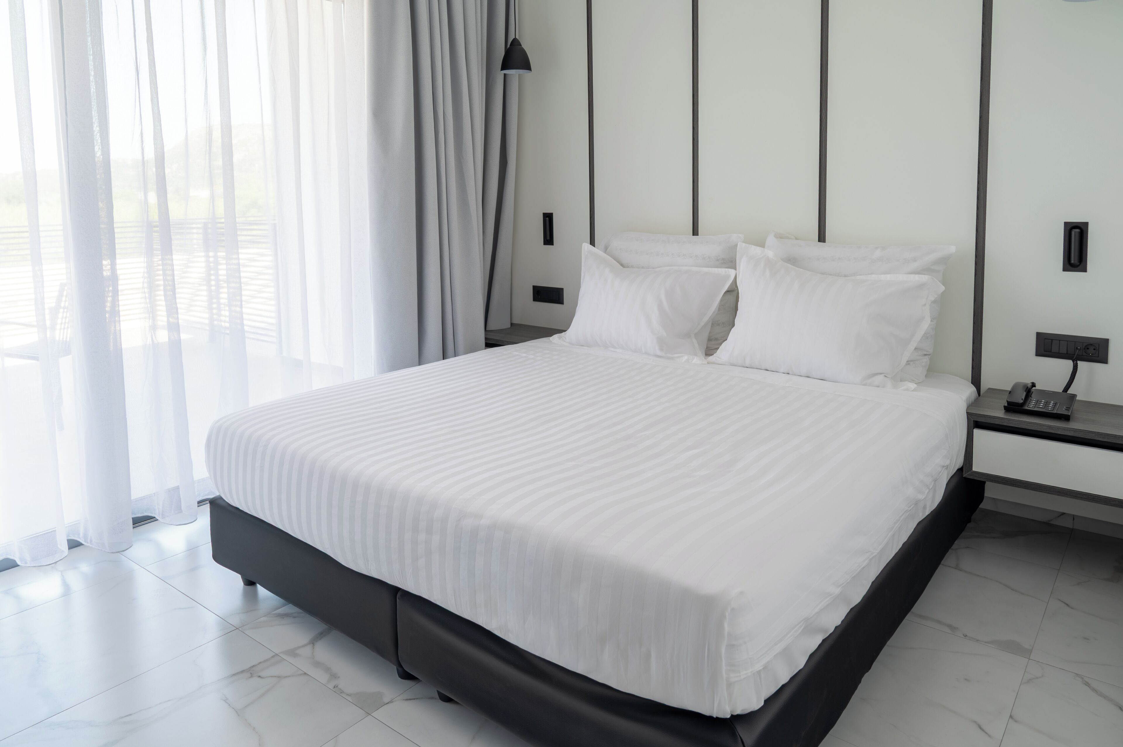 A modern hotel room with a large bed, a nightstand, a lamp, and sheer curtains.