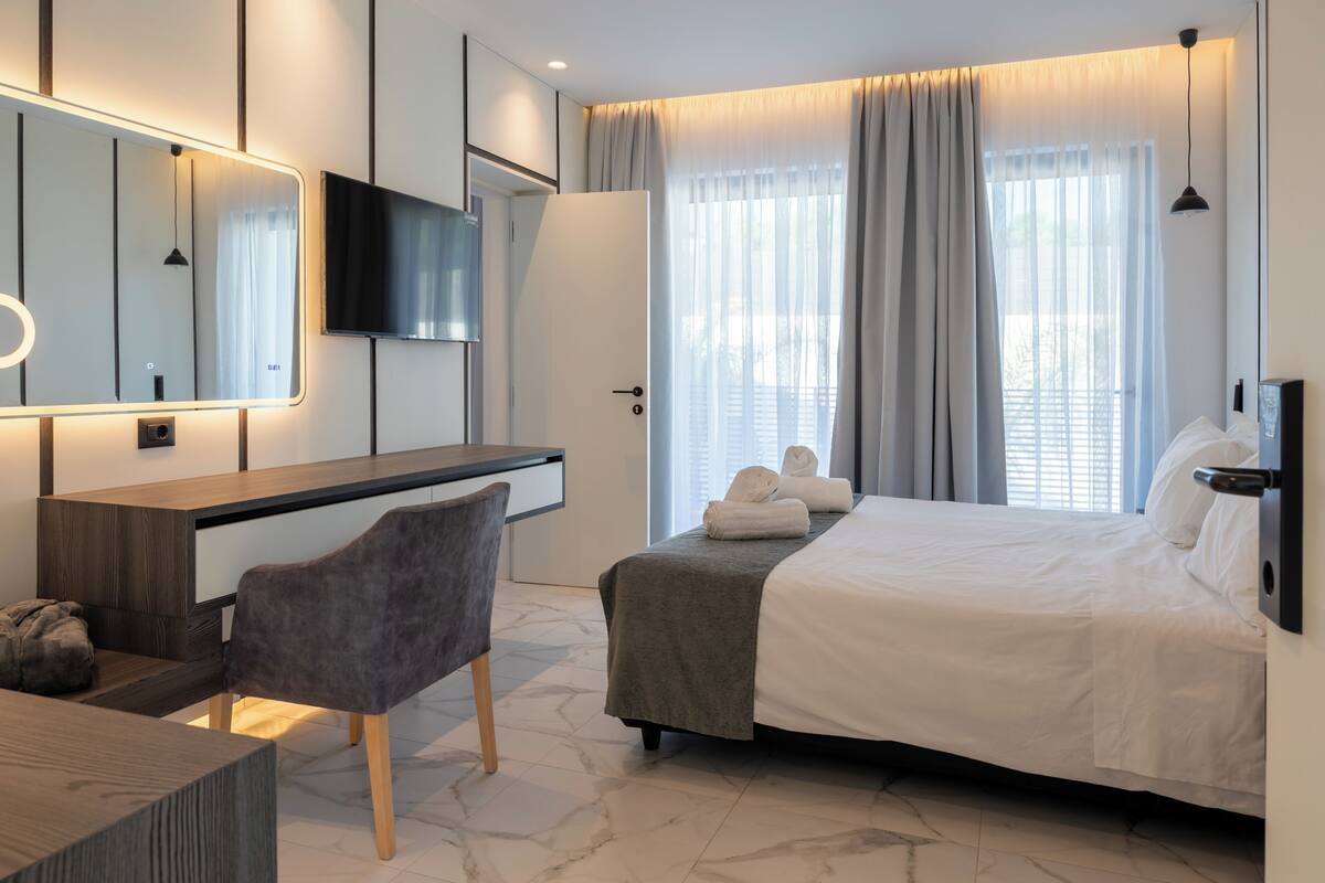 A modern hotel room with a bed, a desk, a chair, a TV, and a large window with curtains.