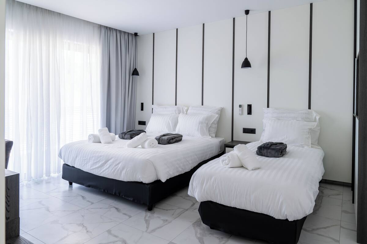 A modern hotel room with two beds, a large window with curtains, a marble floor, and a minimalist design.