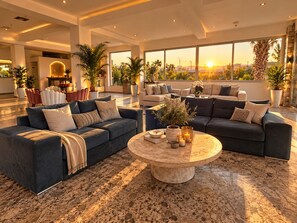 A spacious living area with a large sectional sofa, a round coffee table, and a view of the sunset through large windows. - Oros Paradise Hotel (Rhodes)