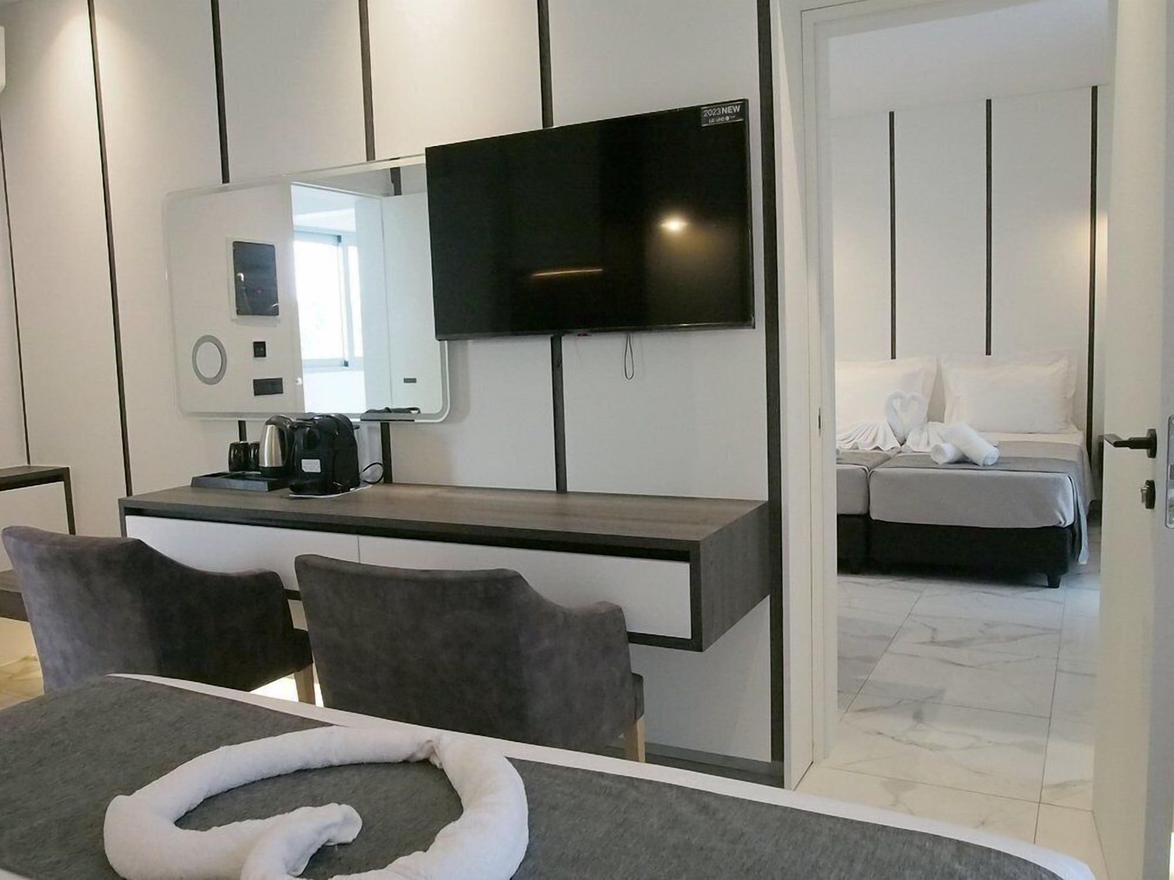 Una habitación de hotel moderna con una televisión de pantalla plana, un escritorio con cafetera, una cama con edredón blanco y un baño visible a través de una puerta abierta.