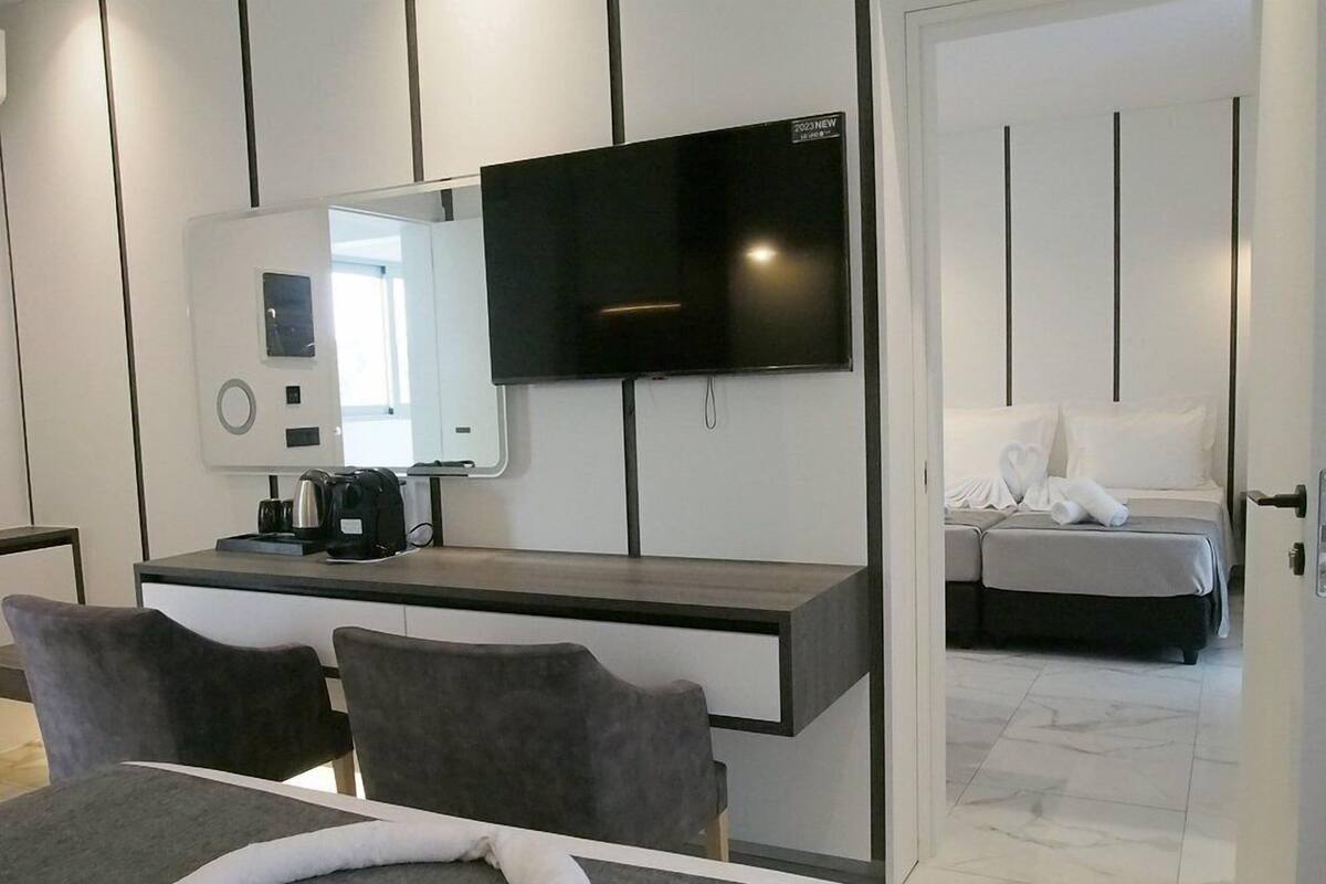 A modern hotel room with a flat-screen TV, a desk with a coffee maker, a bed with a white comforter, and a bathroom visible through an open door.